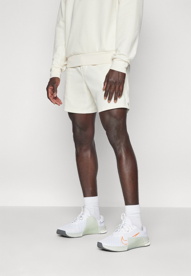 Zapatilla blanca con suela gris, panel lateral verde y detalle naranja. Combinada con calcetines blancos y pantalones cortos y sudadera color crema. Textura suave.