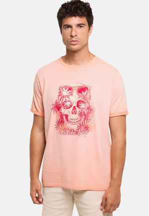 Hombre con una camiseta rosa claro que presenta una calavera roja con gafas de sol y palmeras, de pie contra un fondo liso.