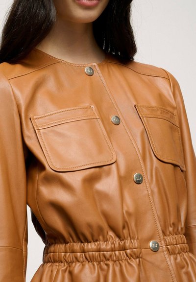 Brown leather jacket with a smooth texture, featuring snap closures, two chest pockets, and a cinched waist design.