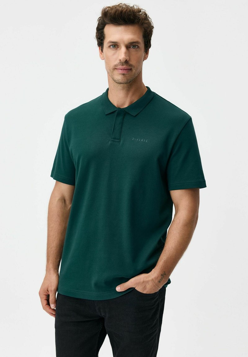 Ciemnozielona koszulka polo z bawełny z kołnierzykiem typu point, krótkimi rękawami i subtelnym logo na piersi. Noszona z czarnymi spodniami.