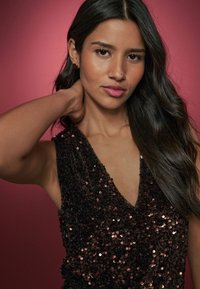 Bezrekawowy top ozdobiony cekinami z głębokim dekoltem w kształcie litery V, charakteryzujący się ciemną paletą kolorów i teksturowaną powierzchnią, na różowym tle.