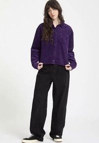 Chemise courte en velours côtelé violet avec boutons et poche poitrine, associée à un pantalon noir ample et des baskets noires. Texture lisse et coupe décontractée.