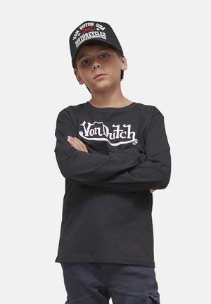 Garçon les bras croisés portant un t-shirt noir à manches longues Von Dutch et une casquette assortie avec des motifs de moto sur un fond uni.