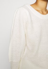 Pull blanc léger en maille avec un motif texturé à mailles ouvertes, un large col et des manches bouffantes. Confectionné en tissu doux.