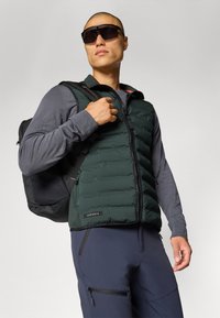 Gilet imbottito verde con chiusura con zip, indossato sopra una maglietta a maniche lunghe grigia. Zaino nero e pantaloni blu navy con tasche con zip. Occhiali da sole scuri.