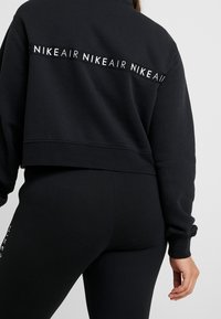 Svart kort sweatshirt med "NIKE AIR"-logotyp i vitt på baksidan, ihop med svarta leggings med en figurnära design och slät textur.