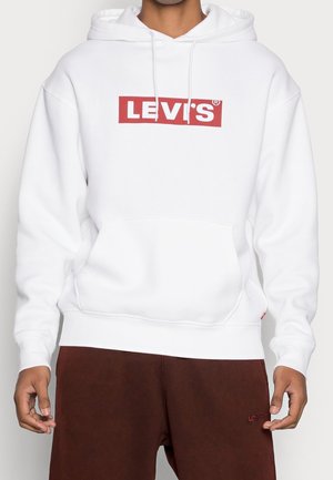 Osoba nosząca białą bluzę z kapturem marki Levi's z czerwonym logo na piersi oraz brązowe spodnie z małym czerwonym logo Levi's na lewej udzie.