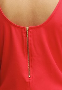 Rotes Oberteil mit niedrigem Rücken, das mit einem goldenen Reißverschluss in der Mitte versehen ist. Glattes Material mit leichtem Glanz und ohne sichtbare Muster.