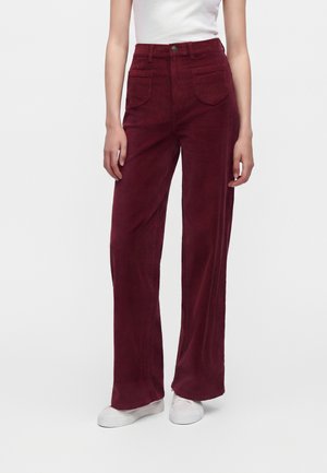 Femme portant un pantalon en velours côtelé bordeaux taille haute à jambes larges avec des poches plaquées devant et des baskets blanches, debout devant un fond uni.