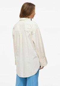 Cămașă oversized bej deschis, cu guler ascuțit, mâneci lungi și tiv curbat. Făcută dintr-un material neted, cu un luciu subtil.