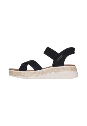 Sandalia negra Skechers tipo slip-on con tiras entrecruzadas, detalle tejido en la entresuela y suela de goma blanca, vista lateral sobre fondo blanco.
