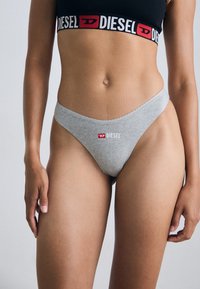 Grijze stringonderbroek met golvende randen, met een rood en wit DIESEL-logo op de voorkant. Het materiaal lijkt zacht en elastisch.