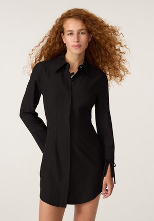 Vrouw met lang krullend rood haar die een aangesloten zwarte lange mouw shirtjurk draagt, staand tegen een effen lichte achtergrond.