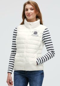 Junge Frau, die eine weiße gesteppte ärmellose Weste über einem langärmligen navyblauen und weißen gestreiften Shirt trägt, steht mit einer Hand in der Tasche.