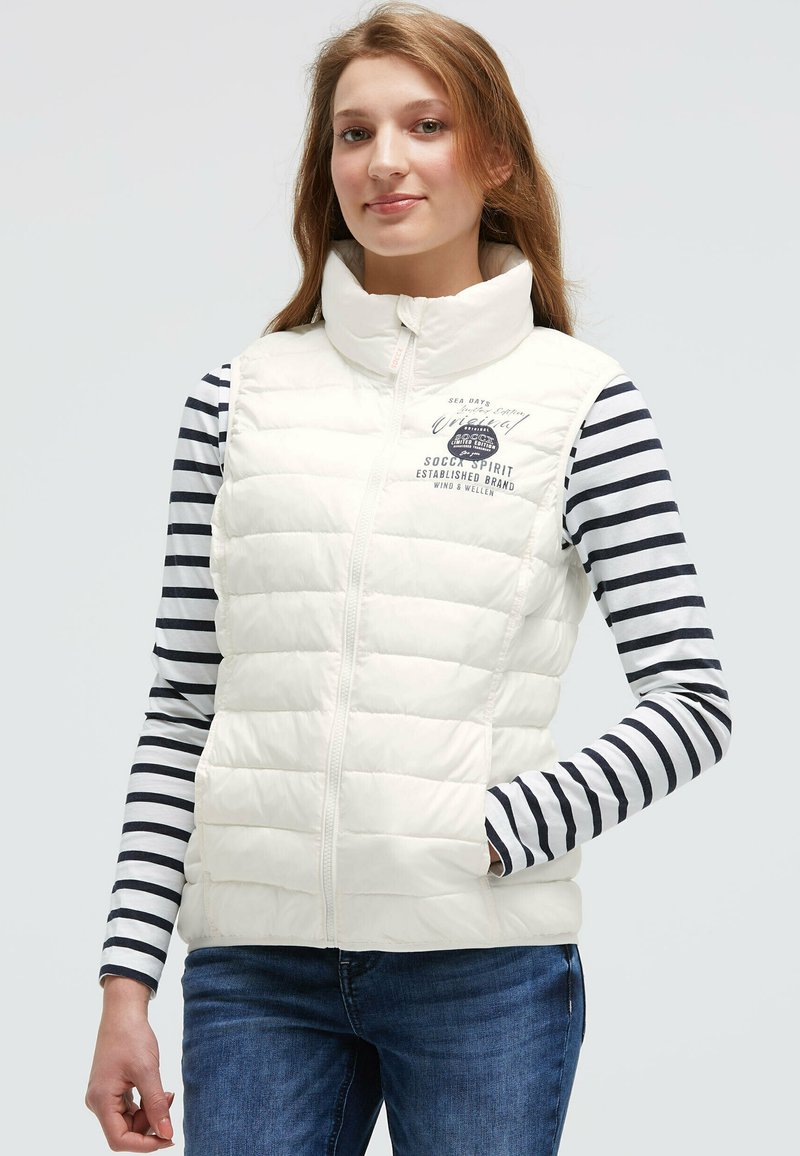 Junge Frau, die eine weiße gesteppte ärmellose Weste über einem langärmligen navyblauen und weißen gestreiften Shirt trägt, steht mit einer Hand in der Tasche.