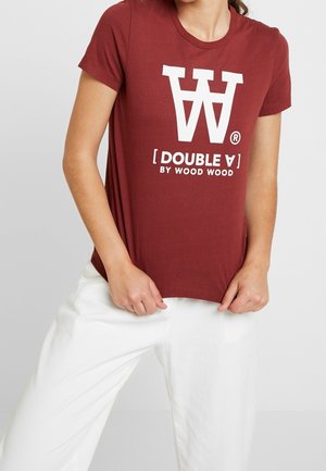 Kobieta nosząca bordową koszulkę z krótkim rękawem z białym napisem "DOUBLE A BY WOOD WOOD" i logo, w połączeniu z luźnymi białymi spodniami.