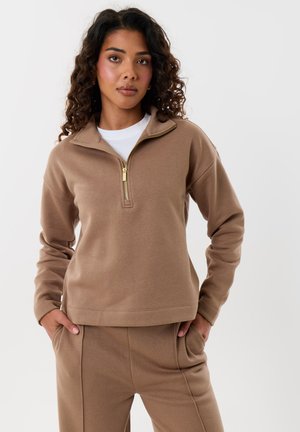 Mujer con cabello rizado que lleva una sudadera beige con cremallera y pantalones a juego, de pie con las manos en los bolsillos frente a un fondo liso.