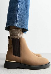 Botas de tobillo de ante en color marrón claro con paneles laterales de elastano y un acento dorado en el tacón. Combinadas con jeans azules desgastados y calcetines beige texturizados.