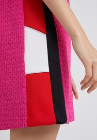 Tissu texturé rose avec un motif ondulé, superposé avec des sections rouges et blanches. Présente un accent latéral noir. Une main est partiellement visible.