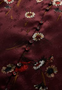 Blusa burdeos con acabado satinado, patrones de flores intrincados y botones decorativos a lo largo de la tapeta delantera.