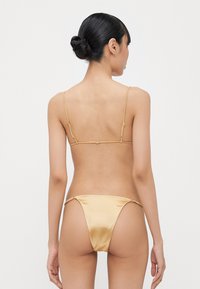 Gold satin bikini with thin adjustable straps and minimal coverage. The back features a delicate clasp and tied accents at the sides.