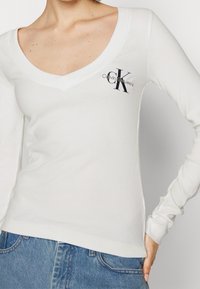 Vit långärmad topp med djup V-ringning, ribbad textur och en liten svart broderad logga på vänster bröst. Matchas med denimjeans.