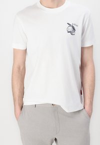 Hombre vestido con una camiseta blanca de manga corta con un pequeño gráfico de limón negro en el lado izquierdo del pecho, ligeramente metida en un pantalón gris claro.