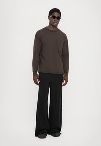 Brown long-sleeve shirt with a chest pocket, paired with loose-fitting black pants. Model wearing dark sunglasses and black shoes.