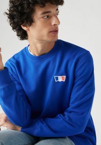Blauwe sweatshirt van katoen met ribbelmanchetten en een ronde hals. Heeft een klein wit, blauw en rood logo op de borst.