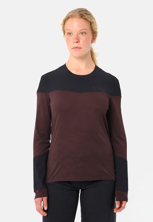 Frau mit lockigem blondem Haar, die ein langärmliges sportliches Shirt in Weinrot und Schwarz trägt und leicht seitlich auf weißem Hintergrund schaut.