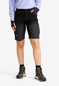 Svart tygshorts med flera fickor med dragkedja, uppvikta nederkanter och en logopatch. Matchas med gröna vandringsskor och en ljuslila topp.