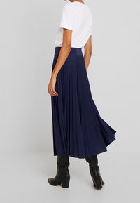 Gonna lunga plissettata blu navy realizzata in tessuto morbido, caratterizzata da un alto punto vita e un design fluido, abbinata a una semplice t-shirt bianca e stivali neri.
