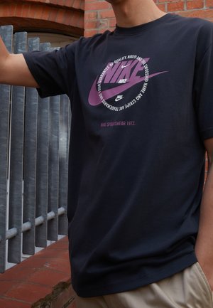 Osoba nosząca czarną koszulkę Nike z fioletowym logo Swoosh i okrągłym białym tekstem, stojąca przy metalowej barierce i ceglanej ścianie.