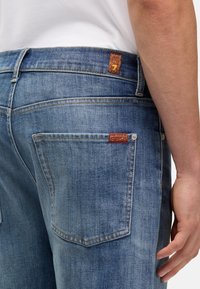 Prześwietlone niebieskie dżinsy z pięcioma kieszeniami, czerwoną i złotą tkana etykietą na pasie oraz kontrastującym przeszyciem wzdłuż szwów.