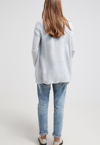 Jasnoszary, dzianinowy kardigan o luźnym kroju, z otwartym plecami i teksturowanym materiałem, w połączeniu z jasnoniebieskimi spodniami typu tapered i mokasynami.