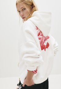 Giovane donna con capelli biondi guarda oltre la spalla indossando una felpa bianca con un grafiche di ciliegie rosa sulla schiena, mano nella tasca.