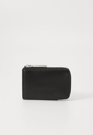 Black leather wallet with a smooth texture, zip closure and embossed "Calvin Klein" logo on the front. Compact rectangular design.