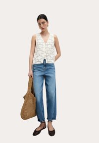 Häkelstrickweste in Weiß mit V-Ausschnitt und Knopfleiste; kombiniert mit hellblauen, hochgeschnittenen Jeans. Das Modell hält eine braune geflochtene Umhängetasche.