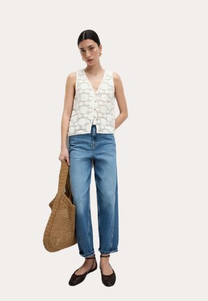Débardeur en crochet blanc avec un col en V et des boutons sur le devant ; associé à un jean taille haute bleu clair. Le mannequin tient un sac fourre-tout tissé marron.