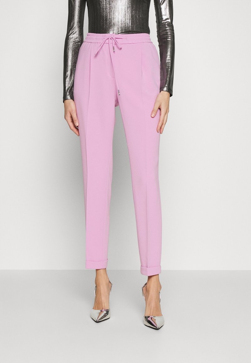 Femme portant un pantalon rose taille haute avec cordon de serrage, ourlets retroussés, et des talons pointus argentés brillants, debout devant un fond uni.