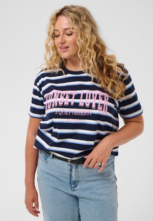 Femme aux longs cheveux blonds bouclés, portant un t-shirt rayé bleu marine avec le texte "SUNSET LOVER" et un jean bleu clair sur un fond uni.