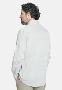 Camisa blanca de manga larga con un patrón texturizado, puños con botones y dobladillo redondeado, llevada sobre pantalones beige claros.