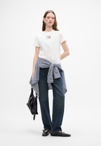 Hvid t-shirt med et lille mønstrede lap, lyseblå og hvidstribet skjorte bundet om taljen, mørkeblå jeans, sort skuldertaske og sorte sko.