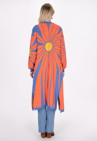 Cardigan lungo in maglia blu, arancione e giallo con motivo a esplosione solare. Presenta maniche lunghe e spacco laterale. Indossato sopra jeans chiari.
