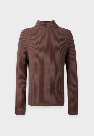 Jeune homme debout portant un pull à col roulé côtelé marron, un pantalon large blanc et des chaussures foncées sur un fond clair uni.