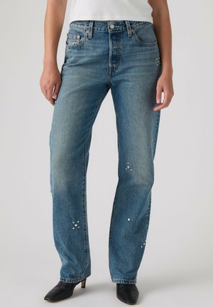 Vrouw draagt blauwe high-waist rechte jeans met kleine strass steentjes en zwarte hakken, gecombineerd met een wit topje.
