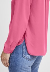 Long-sleeve blouse in bright pink, featuring a smooth texture and subtle shine, paired with light blue denim jeans with faded details.