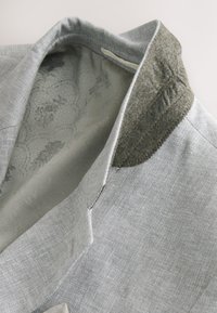Veste en tissu gris clair avec doublure à motif floral et détail de col en feutre gris foncé, plié ouvert.