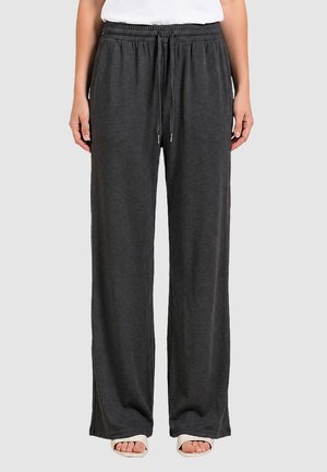 Persona che indossa pantaloni larghi grigio scuro con coulisse e tasche, un top bianco e sandali aperti in punta, in piedi su uno sfondo chiaro e semplice.