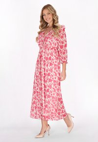 Florales Maxikleid in Hellrosa mit dunkelrosa Blumen, ausgestattet mit einem V-Ausschnitt und Drei-Viertel-Ärmeln. Kombiniert mit beigen High Heels.